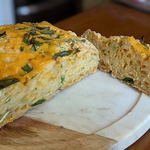 May include: A loaf of cheesy bread with green onions and herbs, sliced in half on a marble cutting board.