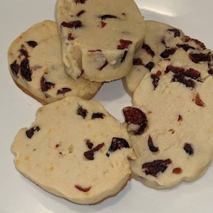 May include: Four shortbread cookies with dried cranberries on a white plate. The cookies are light brown and have a slightly golden hue.