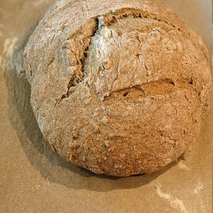 May include: A freshly baked loaf of sourdough bread with a slightly cracked crust. The bread is on a piece of parchment paper.
