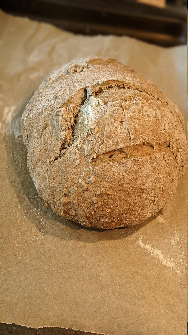 May include: A freshly baked loaf of sourdough bread with a slightly cracked crust. The bread is on a piece of parchment paper.