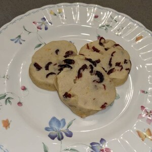May include: Three round, light brown cookies with red cranberries on a white plate with a floral design.