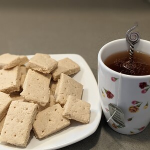 May include: A white plate with a pile of shortbread cookies and a white mug with a floral pattern filled with tea and a metal tea infuser.
