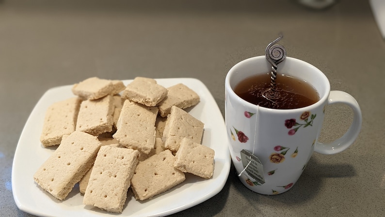 May include: A white plate with a pile of shortbread cookies and a white mug with a floral pattern filled with tea and a metal tea infuser.