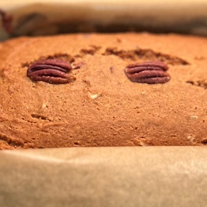 May include: A close-up of a loaf of bread with pecans on top. The bread is in a baking pan lined with parchment paper.