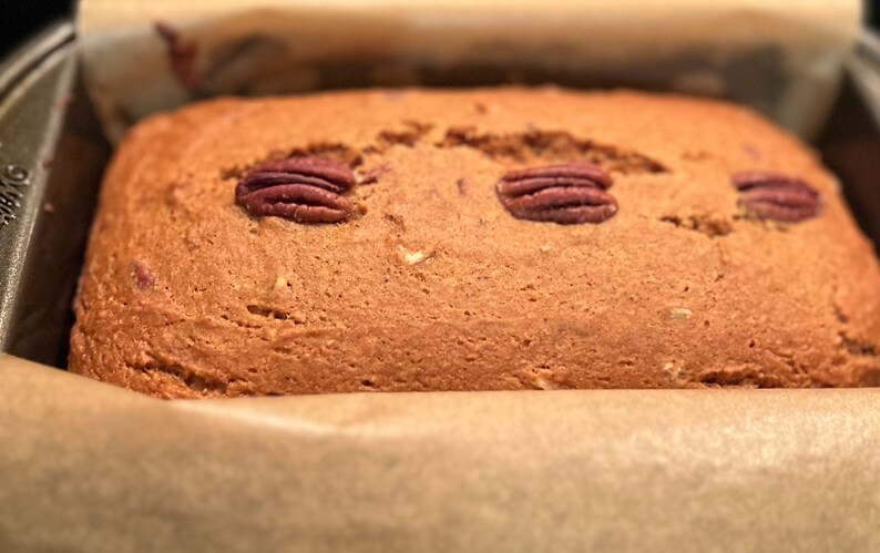 May include: A close-up of a loaf of bread with pecans on top. The bread is in a baking pan lined with parchment paper.