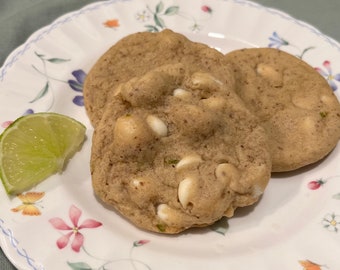May include: Two white chocolate chip cookies on a white plate with a lime wedge. The cookies are soft and chewy, with a light brown color. The lime wedge is green and has a juicy appearance.