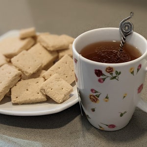 May include: A white mug with a floral pattern filled with tea and a silver spoon. A plate of shortbread cookies sits next to the mug.