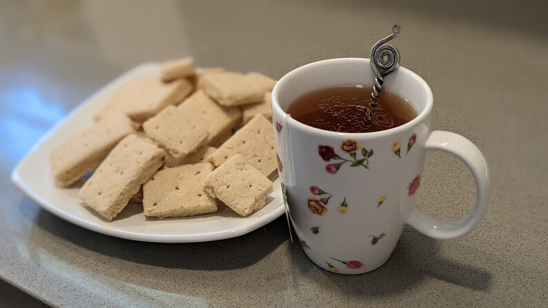 May include: A white mug with a floral pattern filled with tea and a silver spoon. A plate of shortbread cookies sits next to the mug.