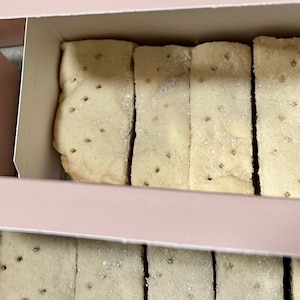 May include: A box of shortbread cookies. The cookies are arranged in two rows, with the top row partially visible. The cookies are light brown and have a slightly crumbly texture. The box is pink and has a white lid.