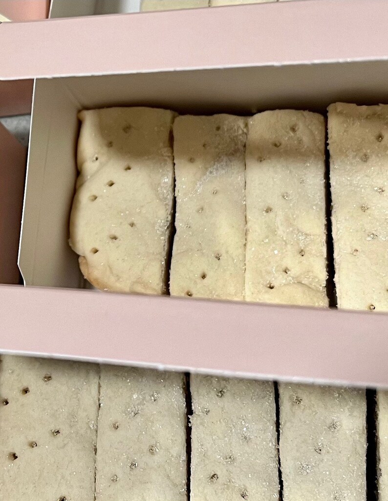 May include: A box of shortbread cookies. The cookies are arranged in two rows, with the top row partially visible. The cookies are light brown and have a slightly crumbly texture. The box is pink and has a white lid.