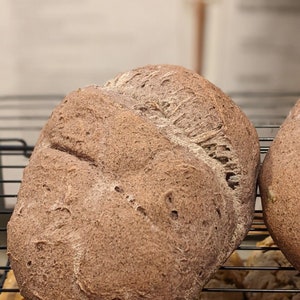 May include: Two dark brown loaves of bread on a black wire rack. The bread has a slightly rough texture and is slightly cracked on the surface.