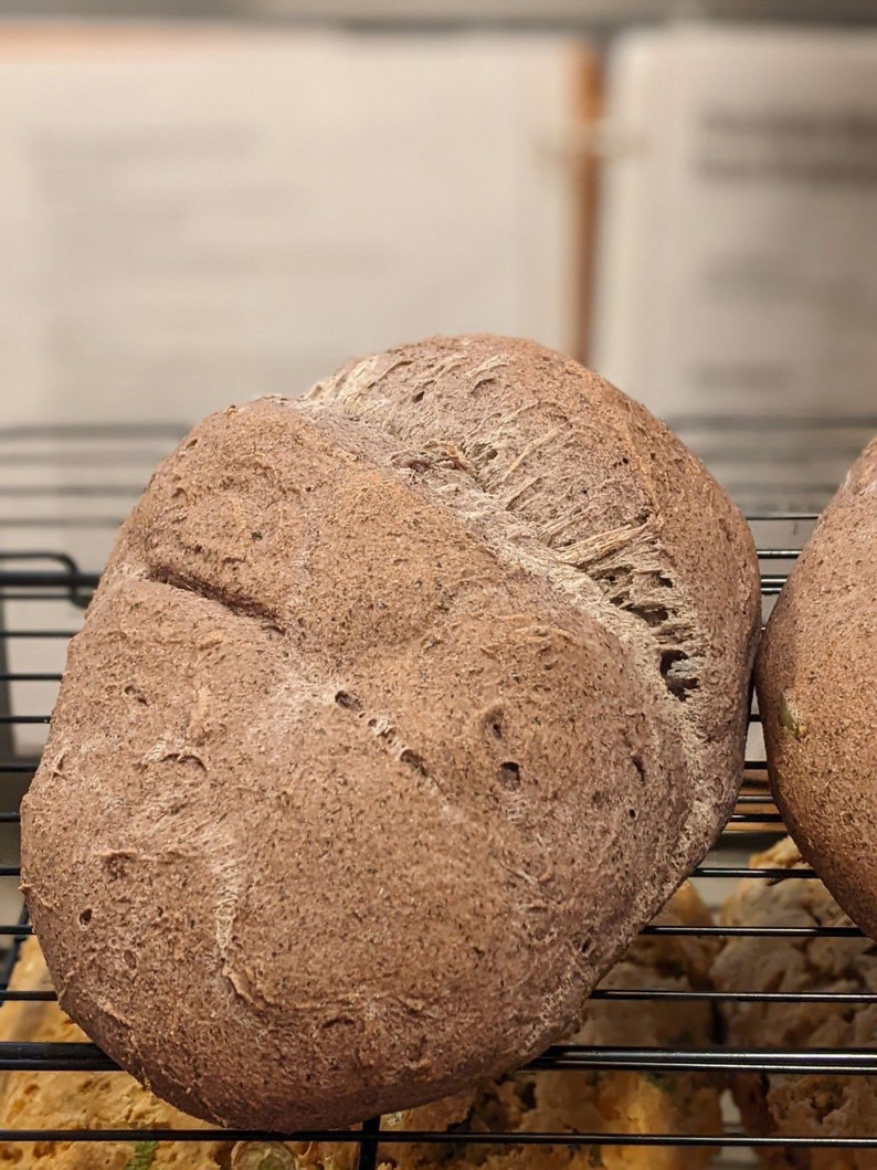 May include: Two dark brown loaves of bread on a black wire rack. The bread has a slightly rough texture and is slightly cracked on the surface.