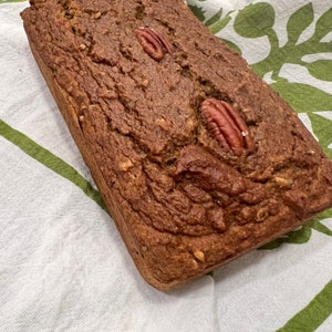 May include: A loaf of brown bread with pecan nuts on top, sitting on a white and green patterned kitchen towel.