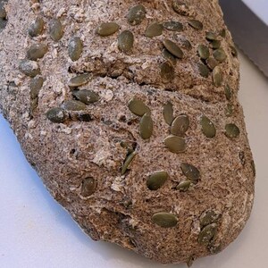 May include: A loaf of dark brown bread with pumpkin seeds on top. The bread is on a white cutting board.