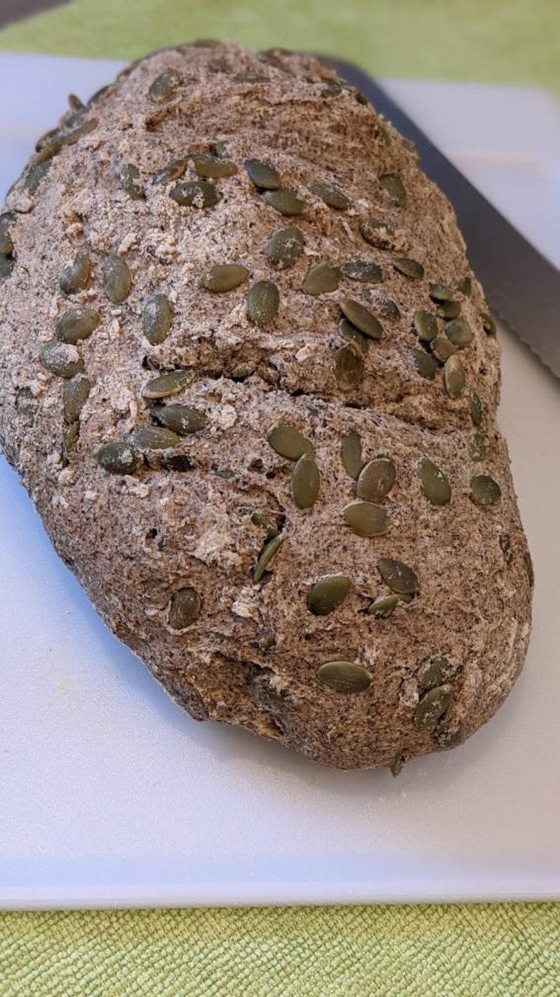 May include: A loaf of dark brown bread with pumpkin seeds on top. The bread is on a white cutting board.