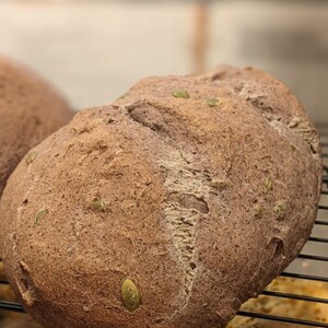May include: A close-up of a dark brown loaf of bread with green seeds on top. The bread is on a black wire rack.