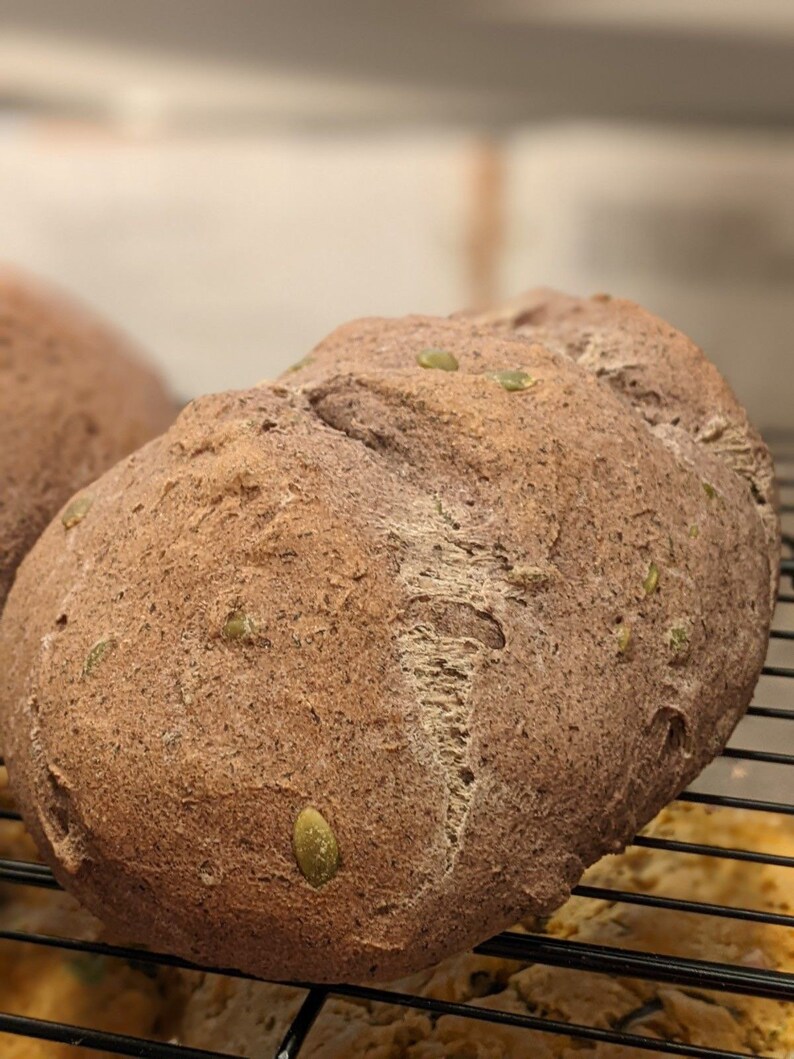 May include: A close-up of a dark brown loaf of bread with green seeds on top. The bread is on a black wire rack.