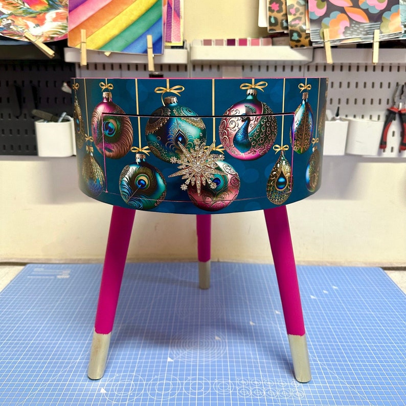 Puede incluir: Una mesa auxiliar redonda con un fondo verde azulado con dise&ntilde;os de adornos navide&ntilde;os. La mesa tiene patas de color rosa brillante con puntas plateadas. Una gran estrella enjoyada est&aacute; centrada en la parte delantera. La mesa tiene un solo caj&oacute;n.