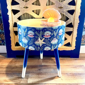 May include: A blue and white side table with a seashell and starfish design. The table has a round top and three legs. A pink seashell with a pearl inside sits on top of the table.