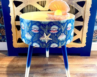 May include: A blue and white side table with a seashell and starfish design. The table has a round top and three legs. A pink seashell with a pearl inside sits on top of the table.