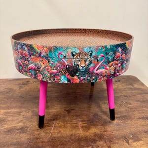 May include: A circular coffee table with a leopard print surface and a colourful wrap featuring a leopard, flamingos, and floral elements. The table has bright pink legs with black tips, creating a bold contrast.
