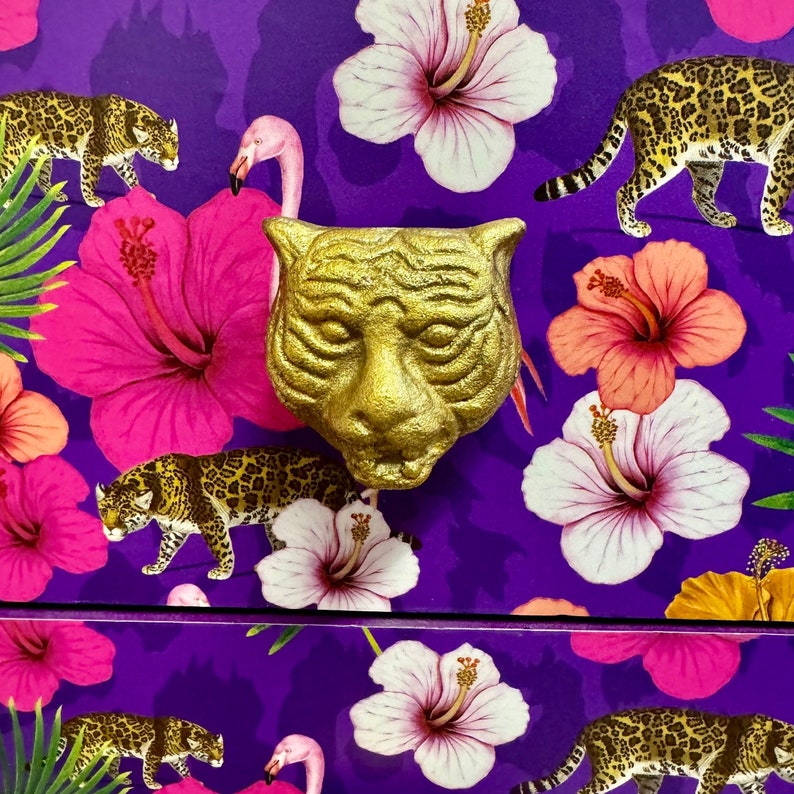 May include: A gold tiger head drawer pull set against a purple background with a tropical design. The design features pink hibiscus flowers, leopards, and flamingos. The pull is a decorative element.