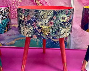May include: A pink side table with a drawer features a colourful print of leopards and jungle foliage. The table has three legs and a round top.