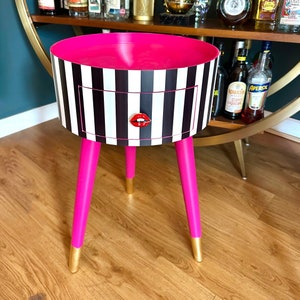 Könnte beinhalten: Ein pink-schwarz gestreifter Beistelltisch mit einer Schublade und drei pinkfarbenen Beinen mit goldenen Akzenten. Die Schublade hat ein rotes Lippen-Design.