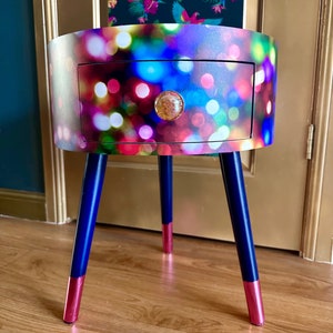May include: A round side table with a drawer and three legs. The table top has a colorful design of blurred lights. The legs are dark blue with a pink accent at the bottom.