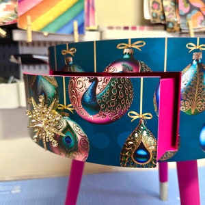 Puede incluir: Peque&ntilde;a mesa auxiliar redonda con fondo verde azulado con adornos navide&ntilde;os dorados. La mesa tiene patas de color rosa brillante con puntas plateadas y un interior rosa. Una estrella dorada adorna el lateral.