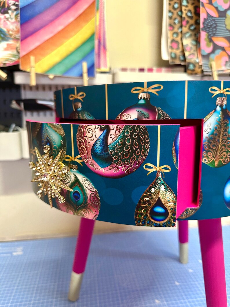 Puede incluir: Peque&ntilde;a mesa auxiliar redonda con fondo verde azulado con adornos navide&ntilde;os dorados. La mesa tiene patas de color rosa brillante con puntas plateadas y un interior rosa. Una estrella dorada adorna el lateral.