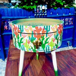 May include: A round side table with a white base and green ombre legs. The table top is covered in a white and green cactus print with pink and orange accents. The table has a single drawer with a gold cactus pull.
