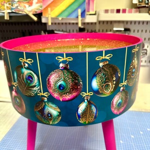 Puede incluir: Una mesa decorativa redonda de color verde azulado con patas rosas y un interior con purpurina dorada. El exterior de la mesa presenta adornos navide&ntilde;os colgantes con dise&ntilde;os de plumas de pavo real. Las patas miden 30 cm de alto.