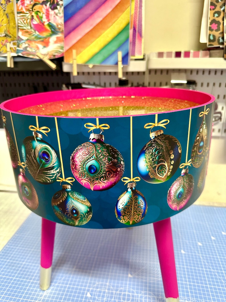 Puede incluir: Una mesa decorativa redonda de color verde azulado con patas rosas y un interior con purpurina dorada. El exterior de la mesa presenta adornos navide&ntilde;os colgantes con dise&ntilde;os de plumas de pavo real. Las patas miden 30 cm de alto.