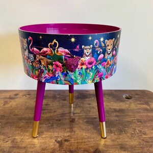 May include: A round side table with a drawer featuring a colorful print of pink flamingos, leopards, and tropical foliage on a dark blue background. The table has three legs with gold tips and a magenta base.