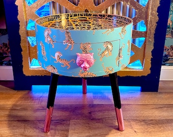 May include: A small, round side table with a drawer. The table is painted a light blue colour and features a tiger print design. The drawer pull is a pink tiger head.