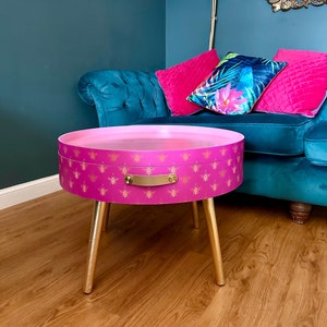 May include: A pink round side table with gold legs and a gold handle. The table top has a pattern of gold bees on a pink background.