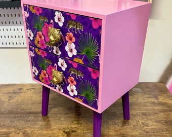 May include: A two-drawer nightstand painted in light pink with purple legs. The drawer fronts feature a tropical pattern with flowers, flamingos, and leopards. Gold-colored tiger head drawer pulls add a decorative touch to this unique piece of furniture.