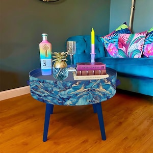 Puede incluir: Una mesa auxiliar azul y dorada con tapa redonda y tres patas. La mesa está decorada con un patrón de mármol. Un libro titulado "American Bar: The Art of Mixing" está encima de la mesa, junto con una botella de licor, una piña y un vaso.