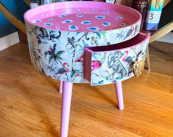 May include: A pink side table with a drawer and three legs. The table is decorated with a tropical print featuring pink flamingos, palm trees, and elephants. The drawer has a leopard print handle.