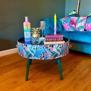 Op de afbeelding: Een ronde bijzettafel met groene poten en een tafelblad met een tropische print. De tafel heeft een zwart oppervlak met een ananas, een fles drank en twee boeken erop. De boeken zijn getiteld "American Bar" en "The Art of Mixing".