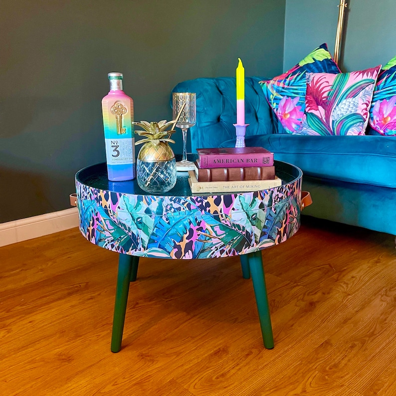 Op de afbeelding: Een ronde bijzettafel met groene poten en een tafelblad met een tropische print. De tafel heeft een zwart oppervlak met een ananas, een fles drank en twee boeken erop. De boeken zijn getiteld "American Bar" en "The Art of Mixing".