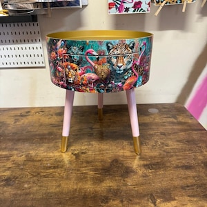 Puede incluir: Una mesa auxiliar redonda con tapa dorada y patas rosas. La mesa está decorada con un estampado colorido de leopardos, flamencos y plantas tropicales.