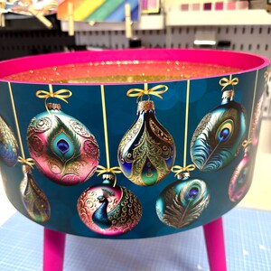 Puede incluir: Una mesa redonda decorativa con un fondo verde azulado con dise&ntilde;os de plumas de pavo real y adornos. La mesa tiene un borde y patas rosas, con purpurina dorada en el interior. Las patas miden unos 30 cm de alto.