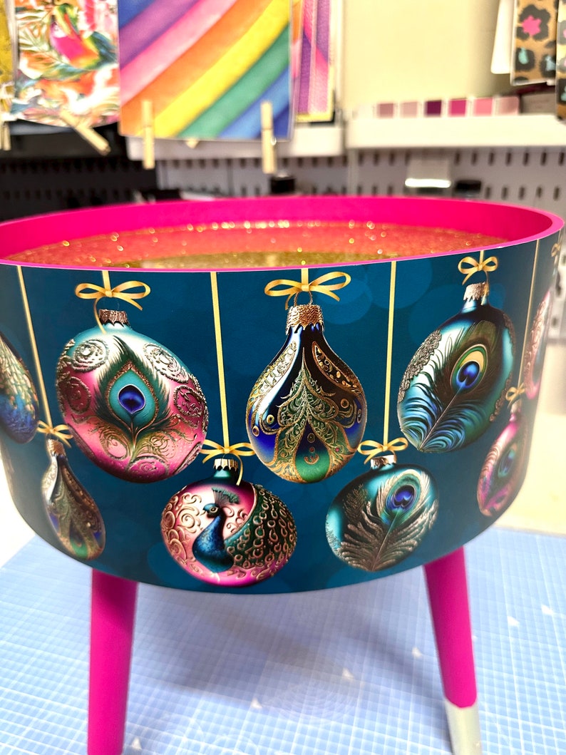 Puede incluir: Una mesa redonda decorativa con un fondo verde azulado con dise&ntilde;os de plumas de pavo real y adornos. La mesa tiene un borde y patas rosas, con purpurina dorada en el interior. Las patas miden unos 30 cm de alto.