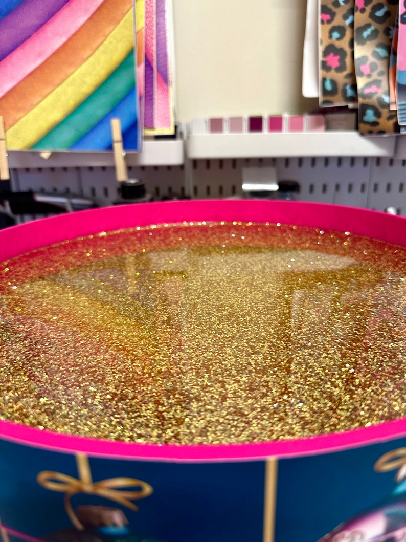 Puede incluir: Un recipiente redondo lleno de purpurina dorada. El recipiente tiene un borde rosa y un exterior azul oscuro con detalles dorados. El fondo incluye papel de colores y otros materiales de manualidades.