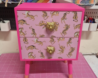 May include: A pink two-drawer dresser with gold tiger print and gold metal drawer pulls. The dresser has short legs.