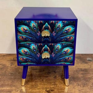 Peut inclure: Une petite table de chevet à deux tiroirs, peinte en bleu profond. Les façades des tiroirs présentent un motif de plumes de paon vibrant, avec des boutons dorés. Les pieds sont bleus avec des embouts dorés.