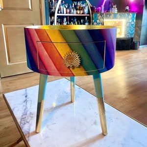 May include: A rainbow-colored side table with a single drawer and gold-painted legs. The drawer has a gold-colored pineapple-shaped knob.