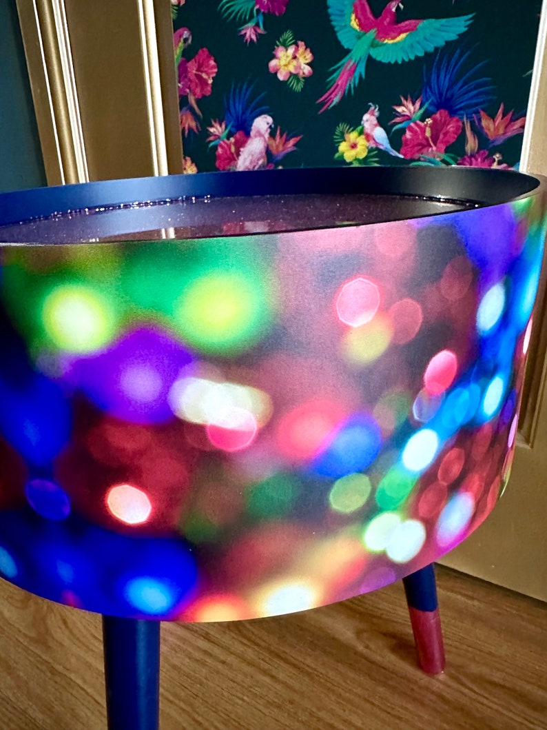 May include: A blue side table with three legs. The table top is covered in a fabric with a blurred out pattern of colorful lights.